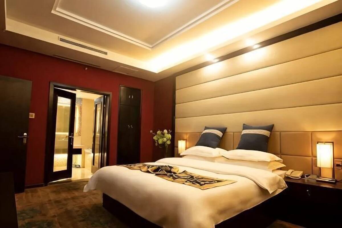A hotel room with a large bed, a bedside table with a lamp, a door, and a wall-mounted light fixture.