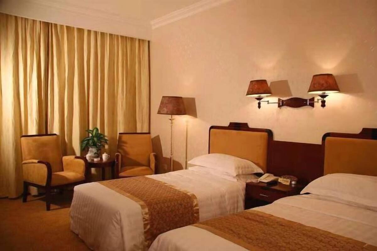 A hotel room with two beds, a sitting area with a chair and a table, a lamp, and a large window with curtains.