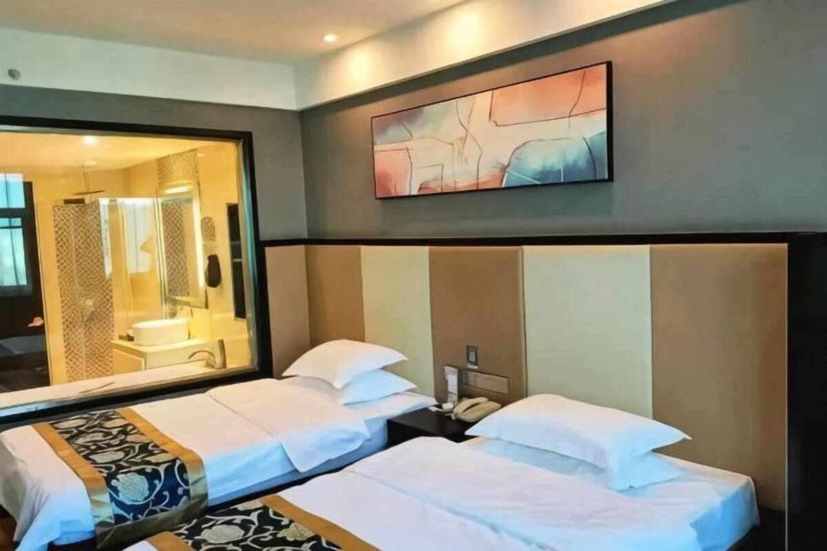A hotel room with two beds, a large mirror, a painting, and a bathroom visible through the glass door.