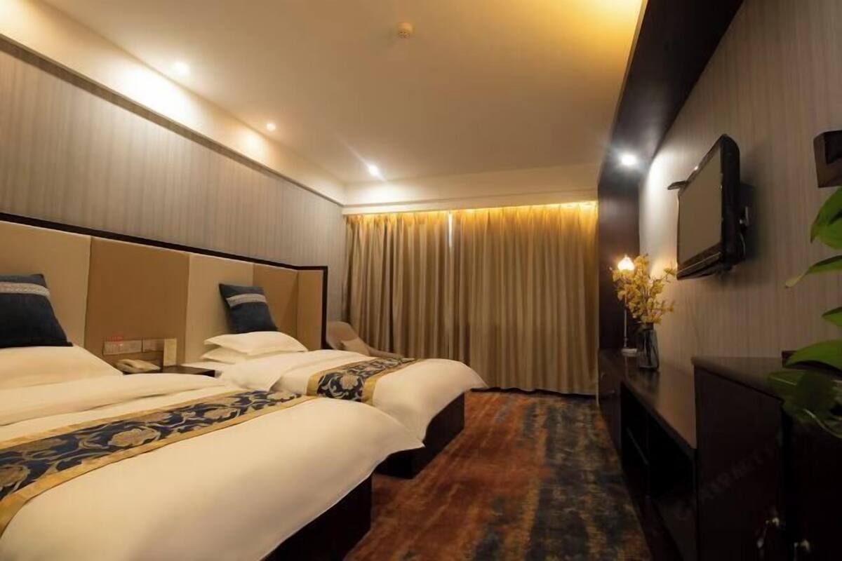 A hotel room with two beds, a flat-screen TV, a desk with a lamp, and a large window with curtains.