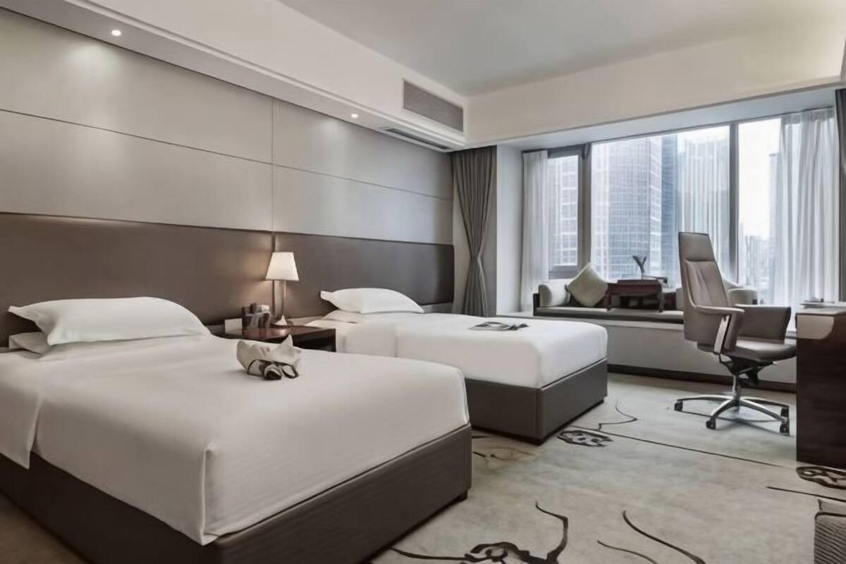A hotel room with two beds, a desk, a chair, and a large window with a city view.