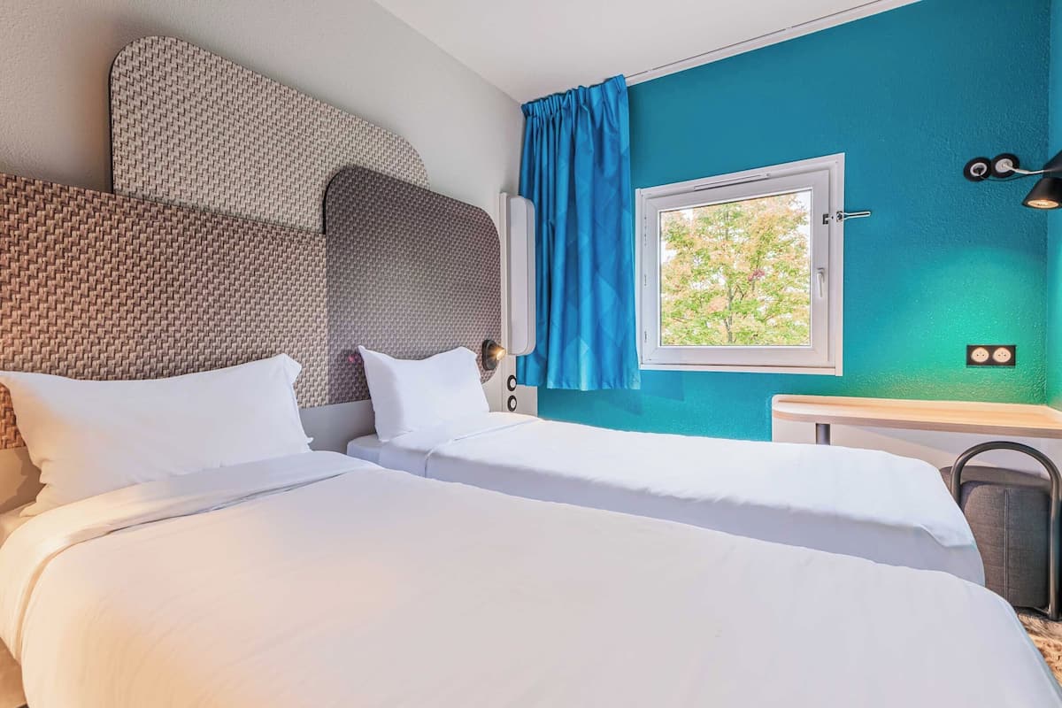 Een hotelkamer met twee bedden, een raam met blauwe gordijnen, een wandlamp en uitzicht op bomen.