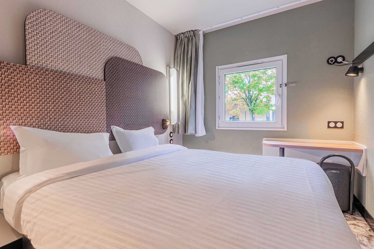 Een hotelkamer met een groot bed, een nachtkastje, een raam met uitzicht op bomen en een wandlamp.