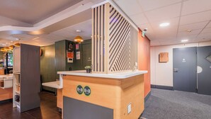 Un comptoir d’accueil avec un revêtement en bois et un logo, un mur gris agrémenté d’un panneau vertical en bois, et une porte avec une indication des numéros de chambre.