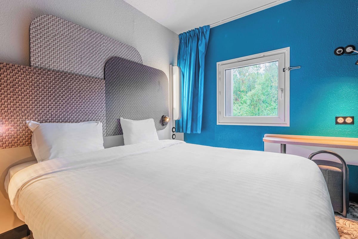 Hotelkamer met een groot bed, een hoofdbord met geweven patroon, een raam met uitzicht op groen en een blauwe wand.