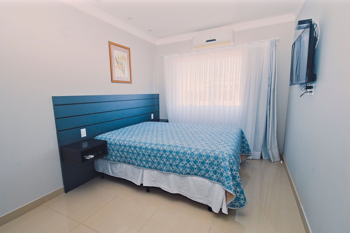 A bedroom with a bed, a nightstand, a wall-mounted TV, and a window with curtains.