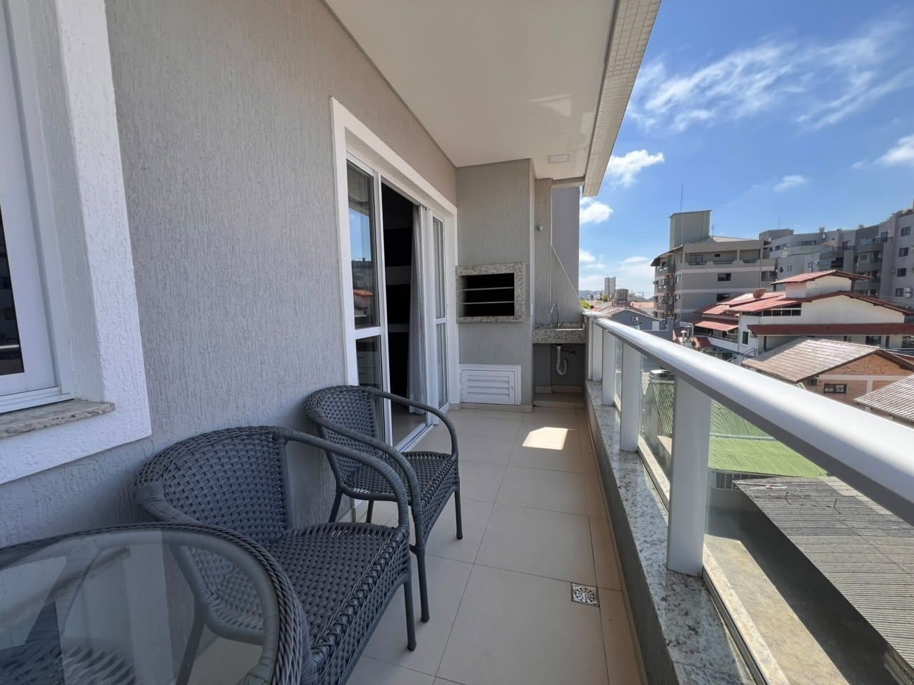 A balcony with two wicker chairs and a small table, a sliding glass door, and a view of neighboring buildings.