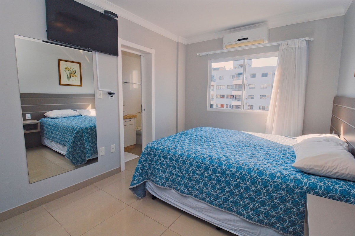 Une chambre à coucher avec un lit, un miroir, une fenêtre avec des rideaux et une vue sur les bâtiments à l’extérieur.