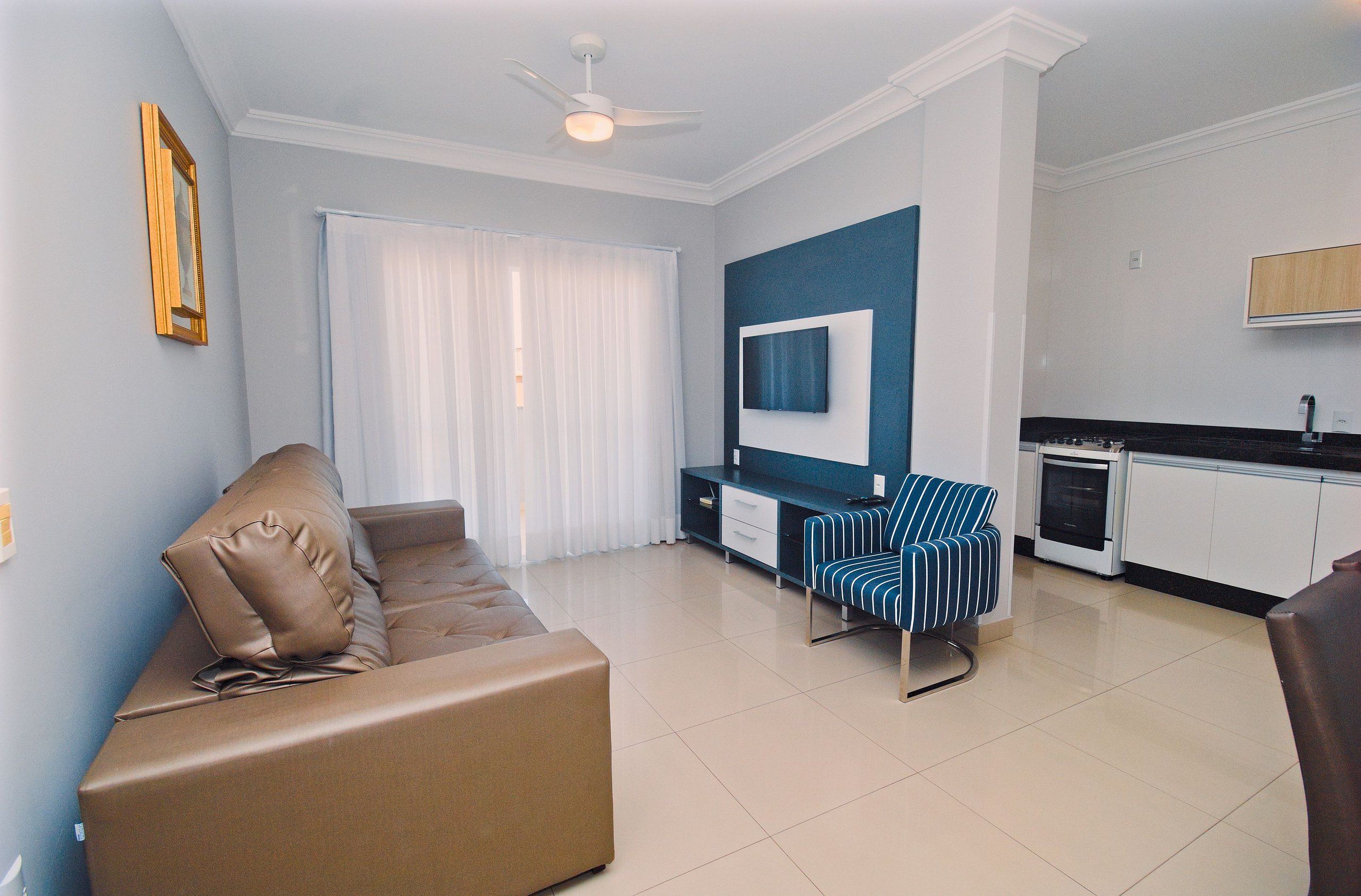 A modern living room with a sofa, a TV mounted on the wall, a dining area, and a kitchen.