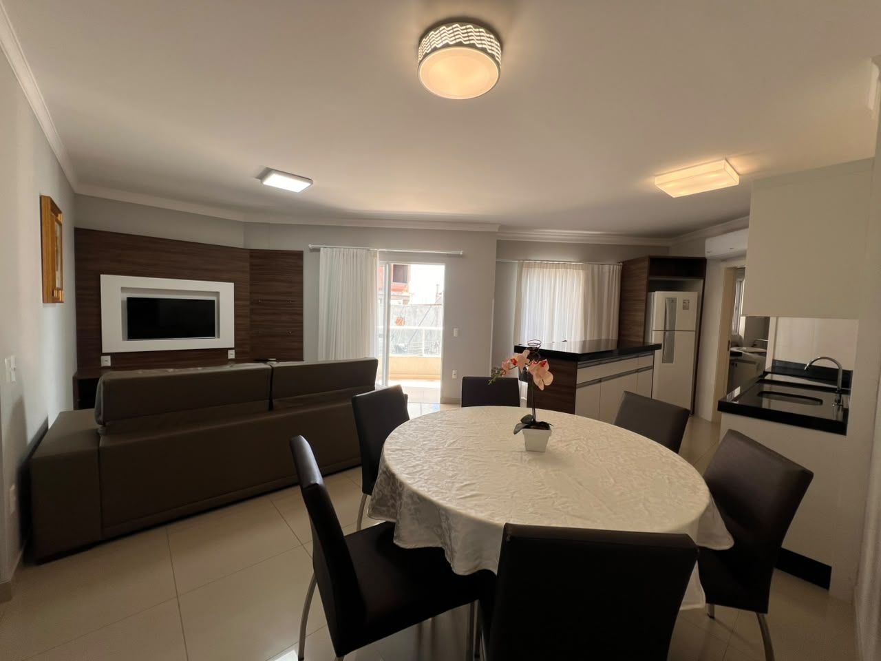 A modern living room with a round dining table, a sofa, a TV mounted on the wall, and a kitchen area with a sink and cabinets.