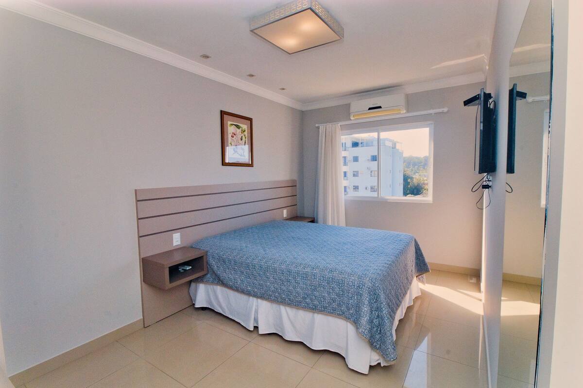 Une chambre à coucher avec un lit, une table de chevet, un tableau encadré au mur et une fenêtre avec des rideaux.
