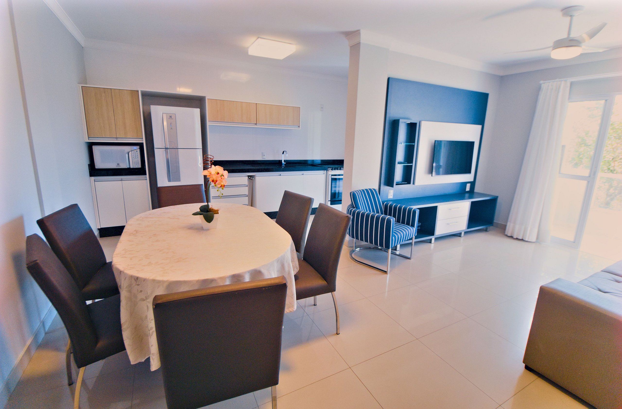 A modern kitchen with a dining area, a TV mounted on the wall, and a large window.