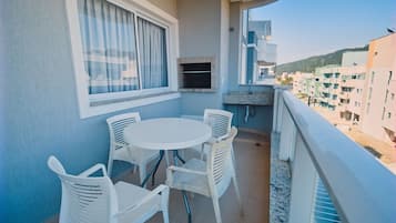 A balcony with a round table and four chairs, a window with curtains, and a view of buildings and hills in the background.
