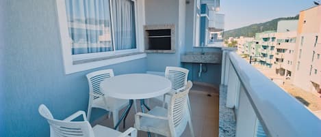 A balcony with a round table and four chairs, a window with curtains, and a view of buildings and hills in the background.