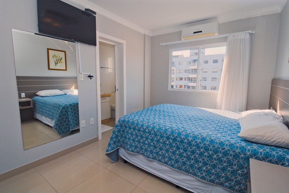 A bedroom with a bed, a mirror, a window with curtains, and a view of buildings outside.