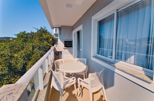 A balcony with a white table and chairs, a window with curtains, and a view of greenery.