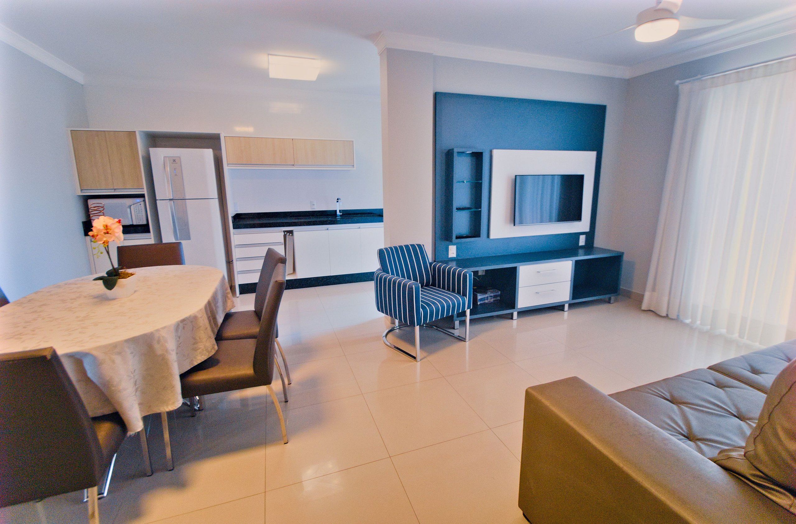 A modern living room with a dining area, a sofa, a TV mounted on the wall, and a kitchen in the background.