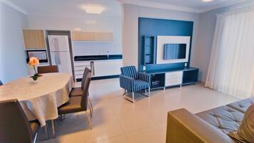 A modern living room with a dining area, a sofa, a TV mounted on the wall, and a kitchen in the background.