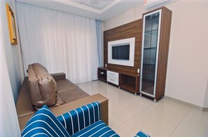 A modern living room with a brown leather sofa, a blue-striped ottoman, a wooden entertainment unit with a TV, and a glass cabinet. -  Residencial Baía dos Golfinhos (Bombinhas)