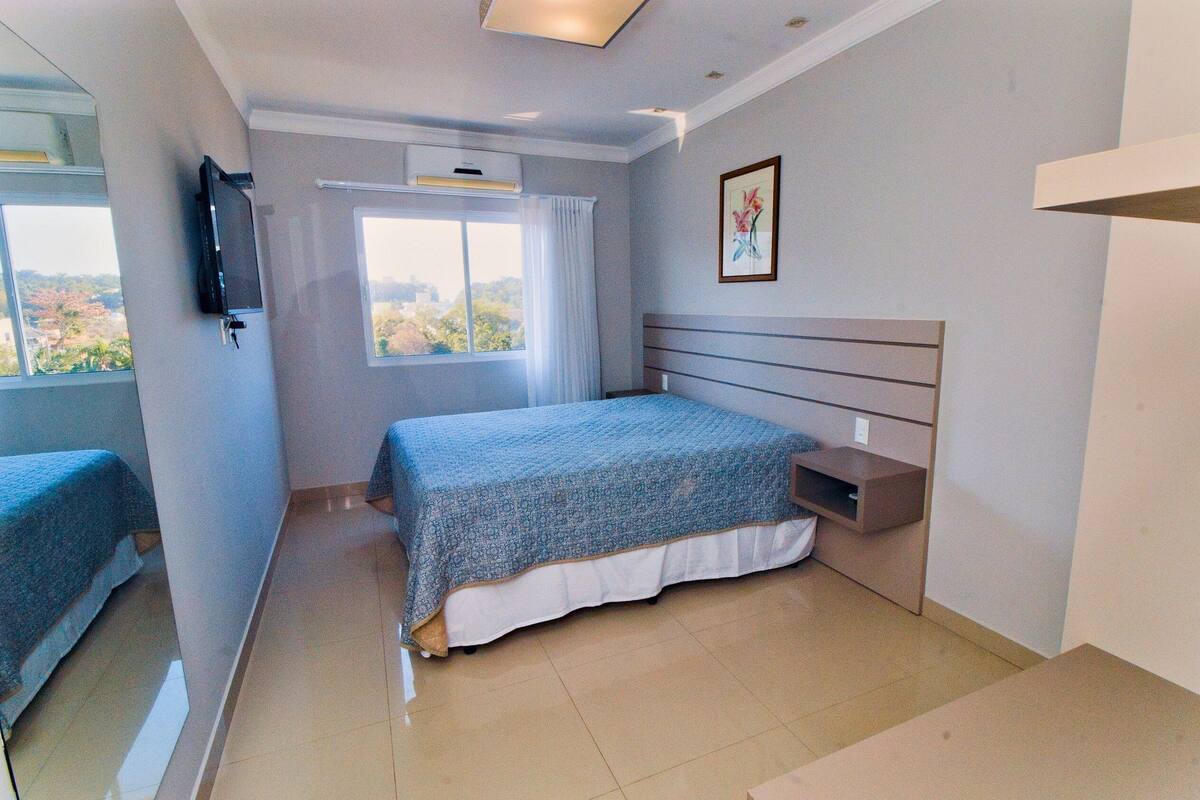 Une chambre à coucher avec un lit, un téléviseur fixé au mur, une fenêtre avec des rideaux et un tableau accroché au mur.
