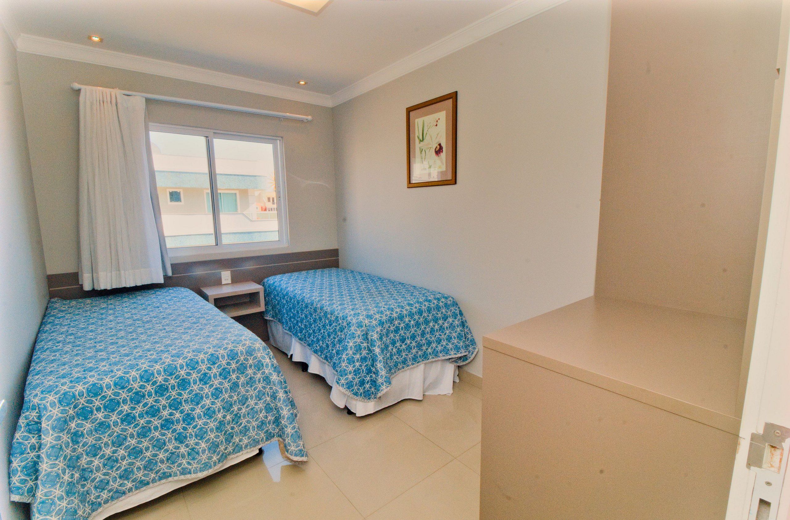 A small, clean room with two single beds, a window with curtains, a small table, and a framed picture on the wall.