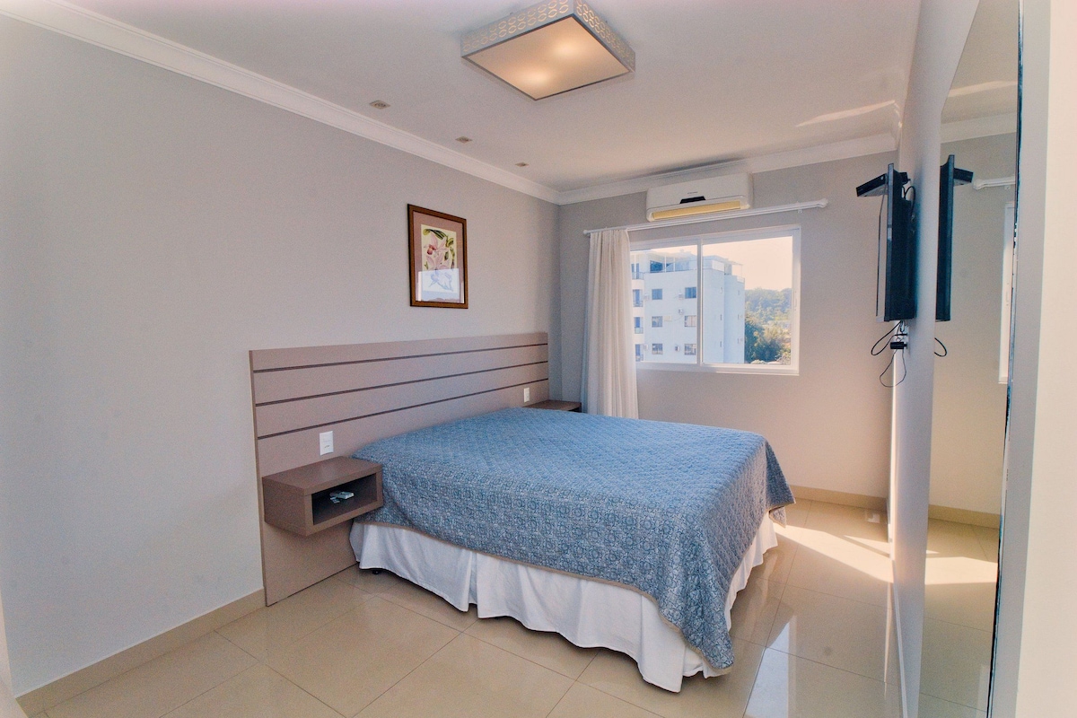 A bedroom with a bed, a nightstand, a framed picture on the wall, and a window with curtains.