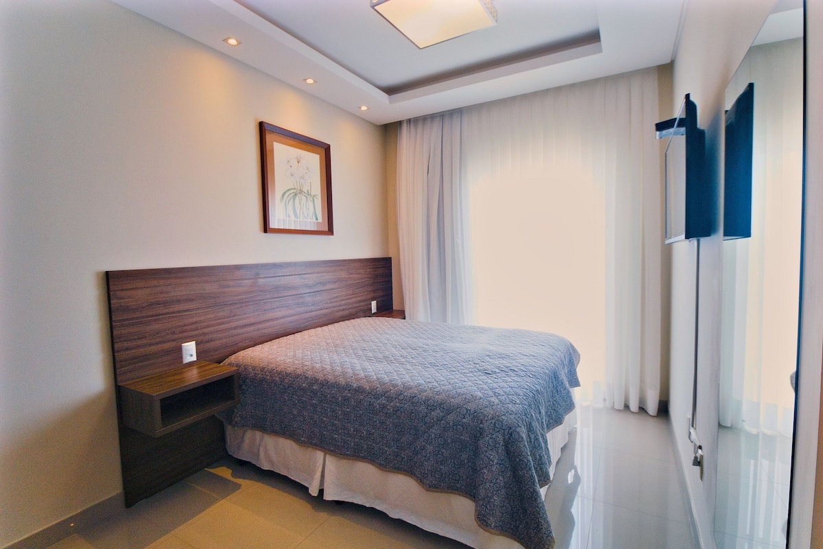 Une chambre à coucher avec une tête de lit en bois, une table de chevet, un lit recouvert d’une couvre-lit à motifs, un tableau encadré au mur et une fenêtre avec des rideaux.