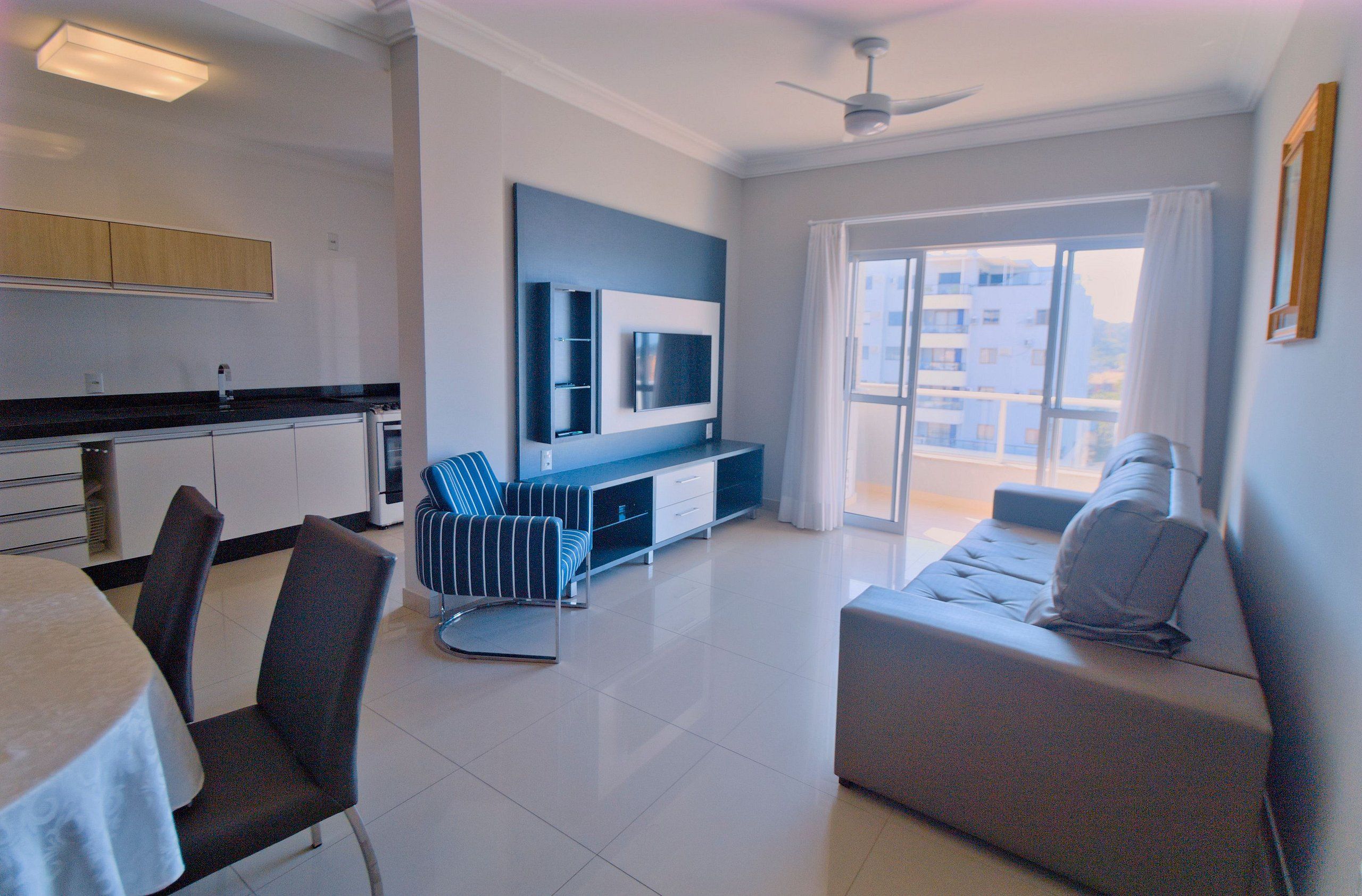 A modern living room with a sofa, a dining table with chairs, a TV mounted on the wall, and a balcony with a view of buildings.