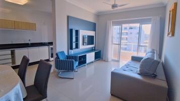 A modern living room with a sofa, a dining table with chairs, a TV mounted on the wall, and a balcony with a view of buildings.