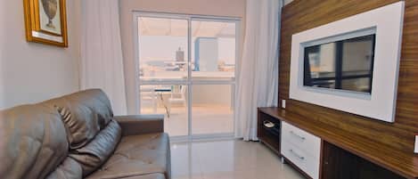 A modern living room with a leather sofa, a TV on a wooden wall, and a sliding glass door leading to a balcony.