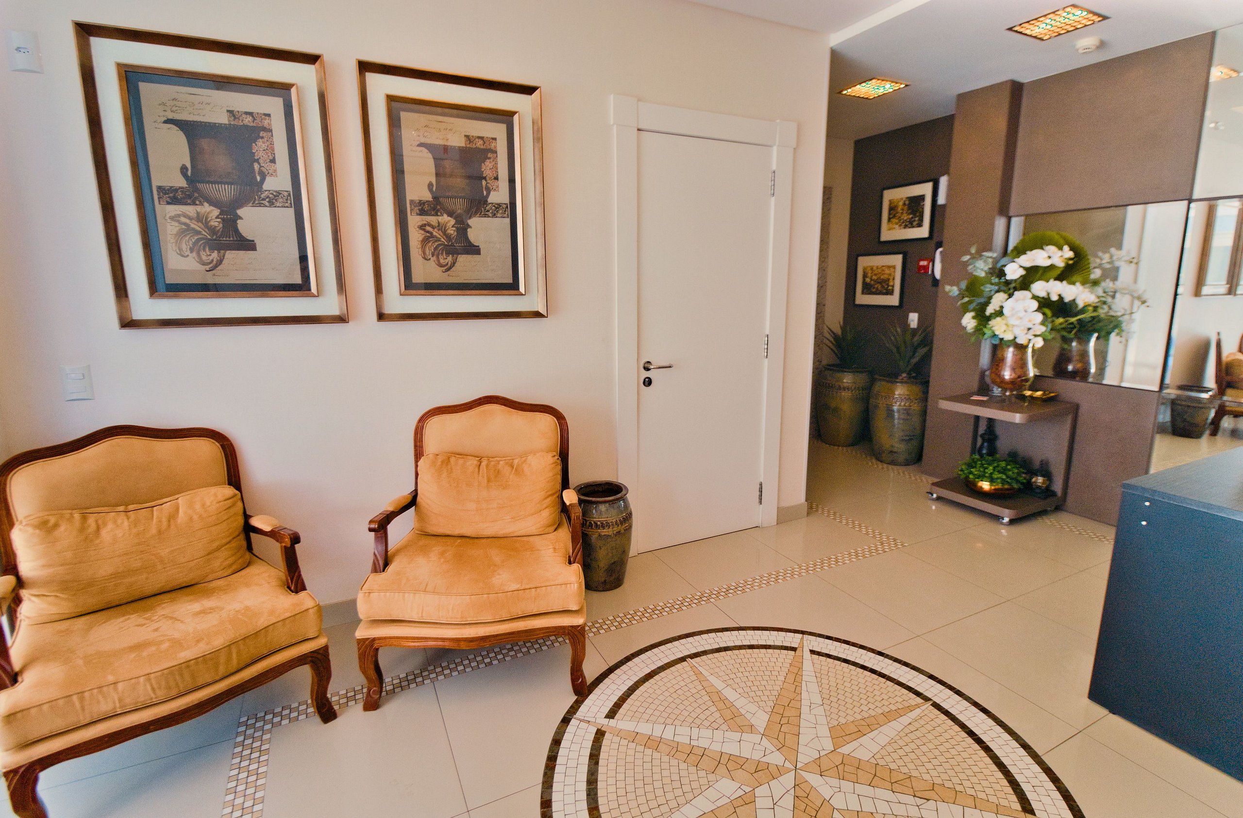 A lobby with two leather armchairs, a tiled floor, framed artwork on the wall, and a reception desk with a vase of flowers.