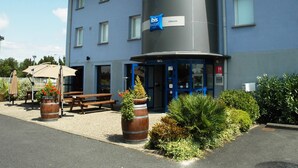 A building with a blue and grey facade, a sign for 'Idis budget' on the entrance, outdoor seating, and planters with flowers. - Ibis Budget Libourne (Libourne)