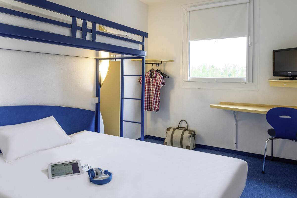 Quarto de hotel com beliche, escrivaninha, cadeira, televisão e janela.