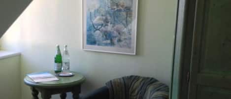 Une pièce avec un lit, une chaise, une petite table sur laquelle se trouve une bouteille, et un tableau accroché au mur.