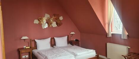 A bedroom with a sloped ceiling, a bed with white bedding, a wooden nightstand, a lamp, and a radiator.