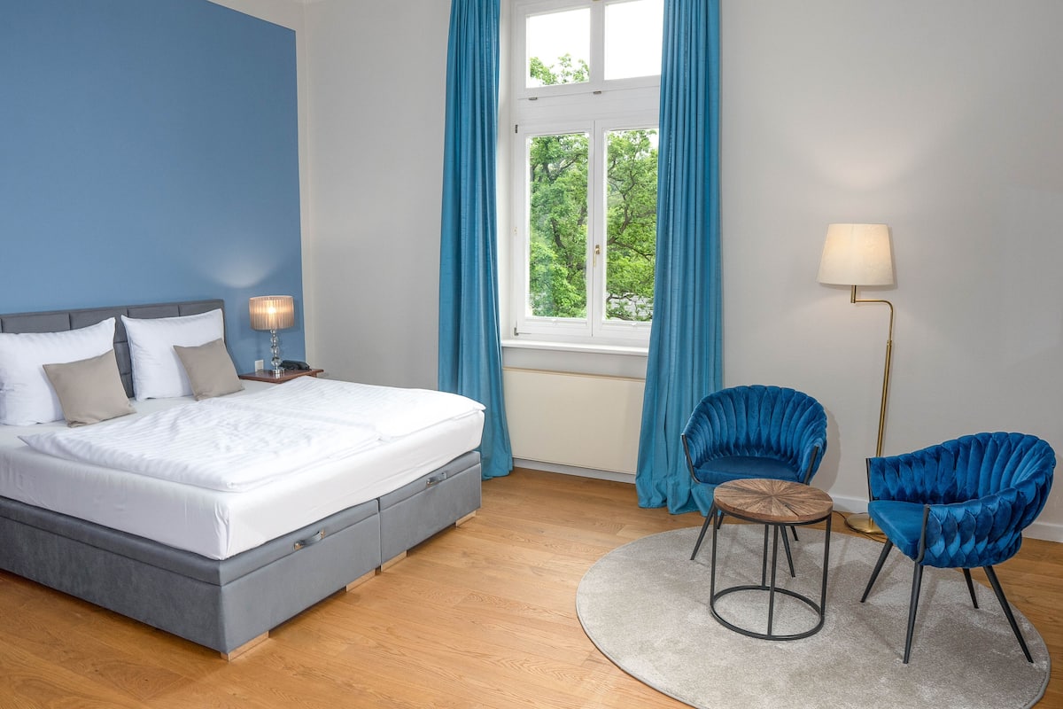 Une chambre à coucher avec un lit, deux chaises, une lampe sur pied et une fenêtre avec des rideaux.