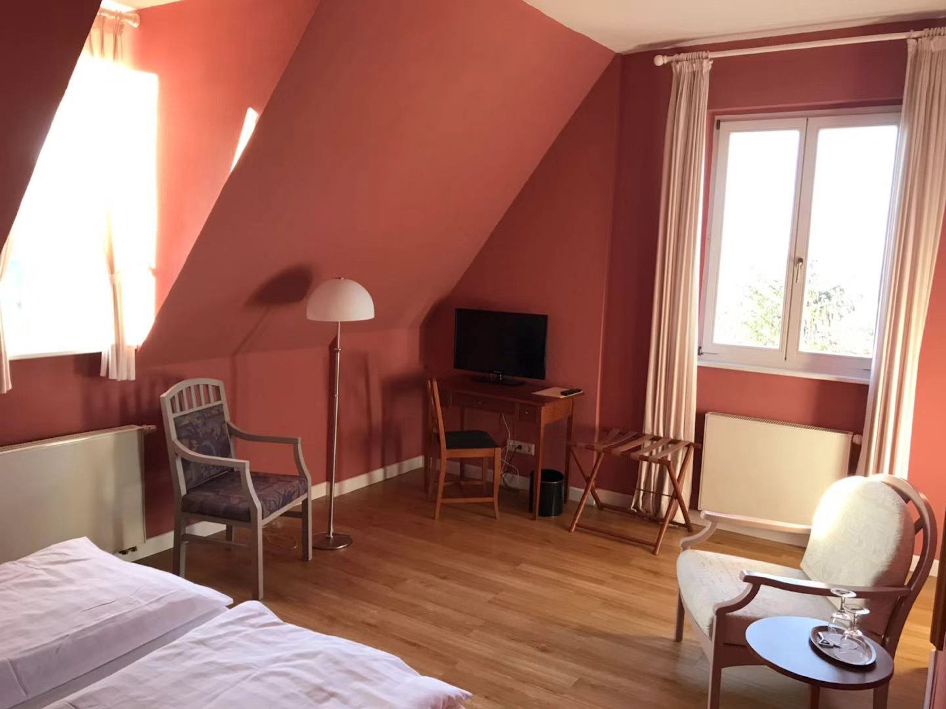 A room with a sloped ceiling, a bed, a desk with a chair, a television, and a window with curtains.
