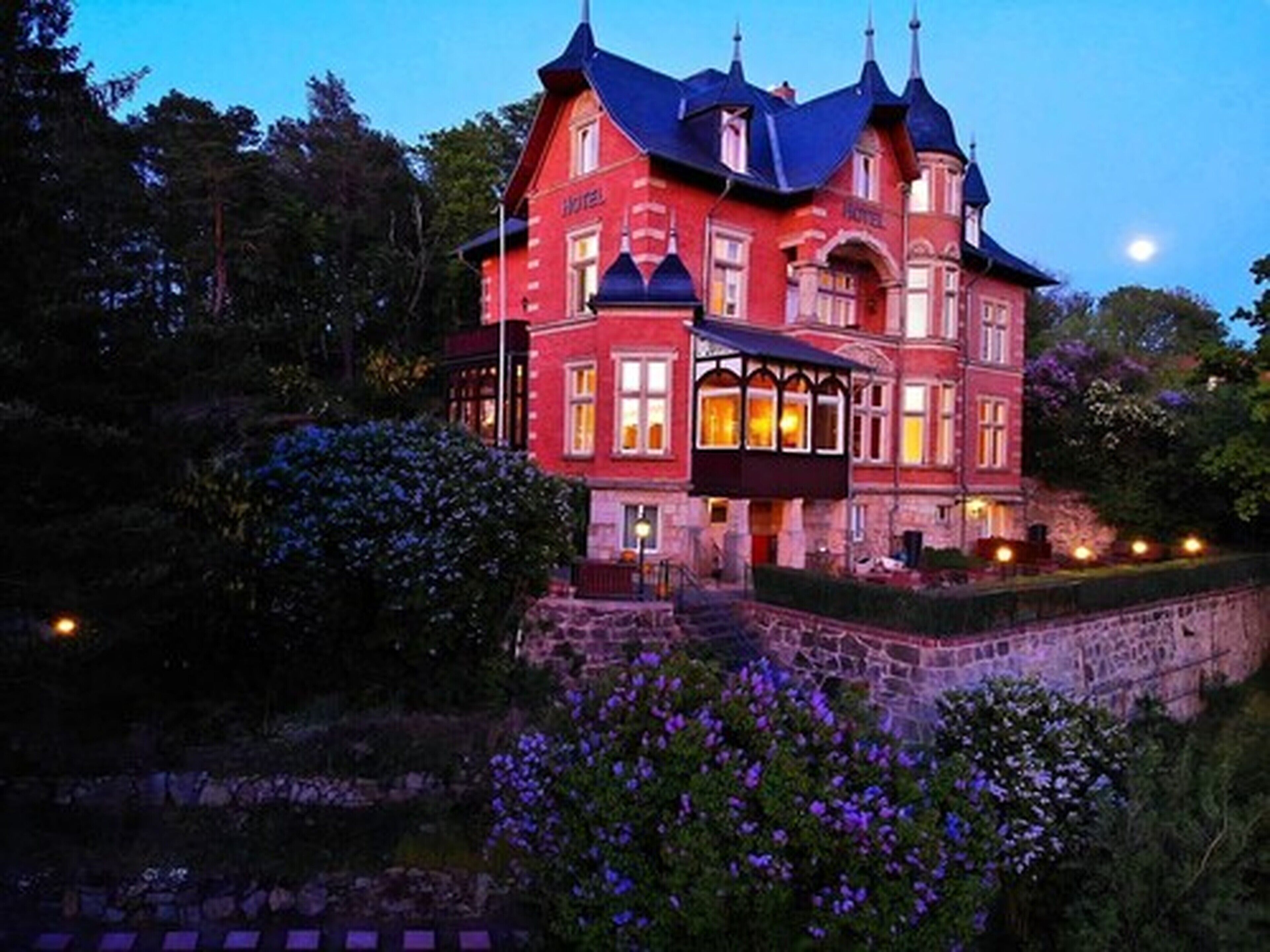 A large, red brick building with a steeply pitched roof and multiple chimneys, likely a hotel, surrounded by lush greenery and flowering plants.