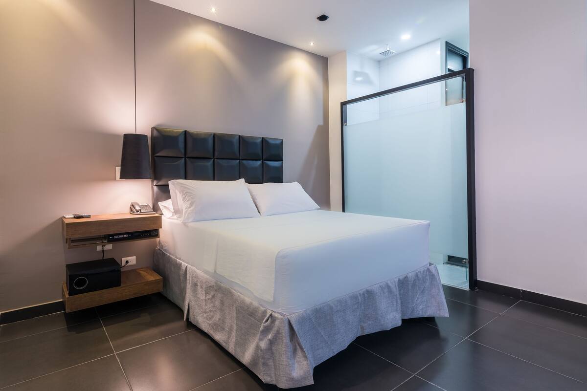A modern hotel room with a large bed, a bedside table with a lamp, and a glass shower enclosure.