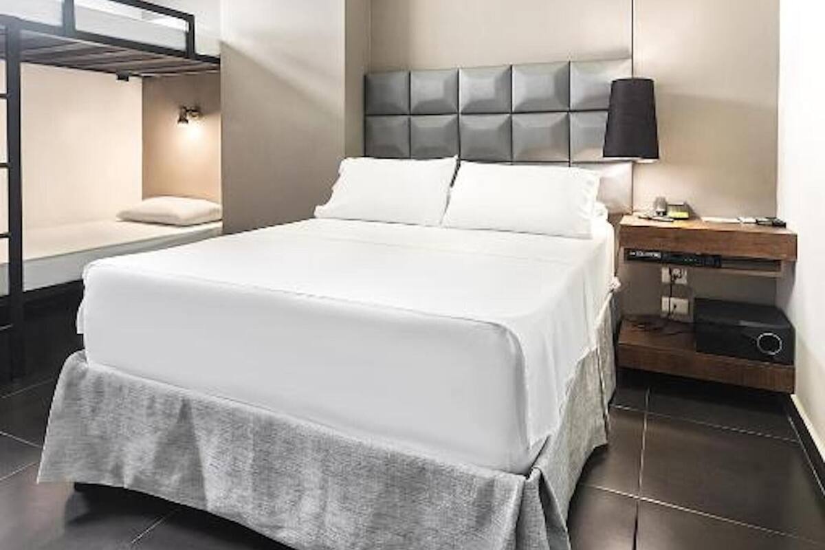 A modern hotel room with a large bed, a bedside table, a wall-mounted shelf, and a bunk bed.