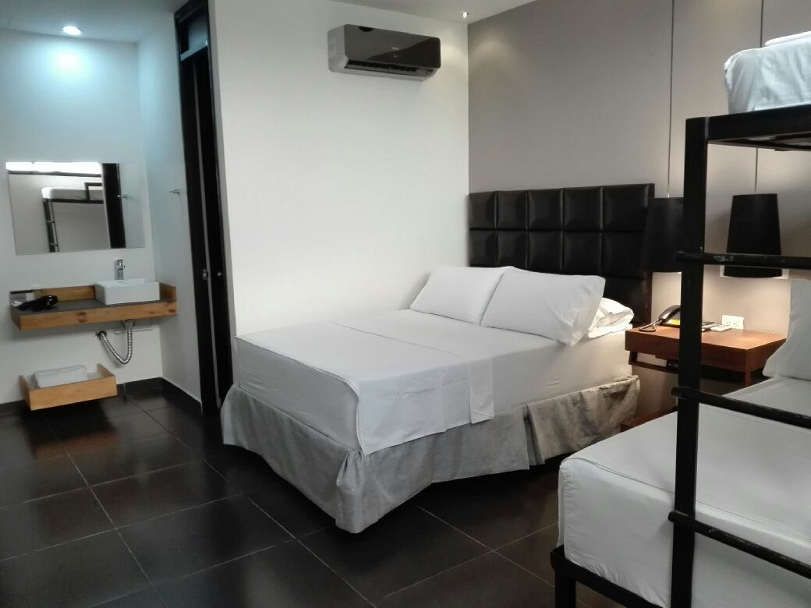 A modern hotel room with a bed, a bedside table with a lamp, a built-in shelf with a mirror, and a black headboard.