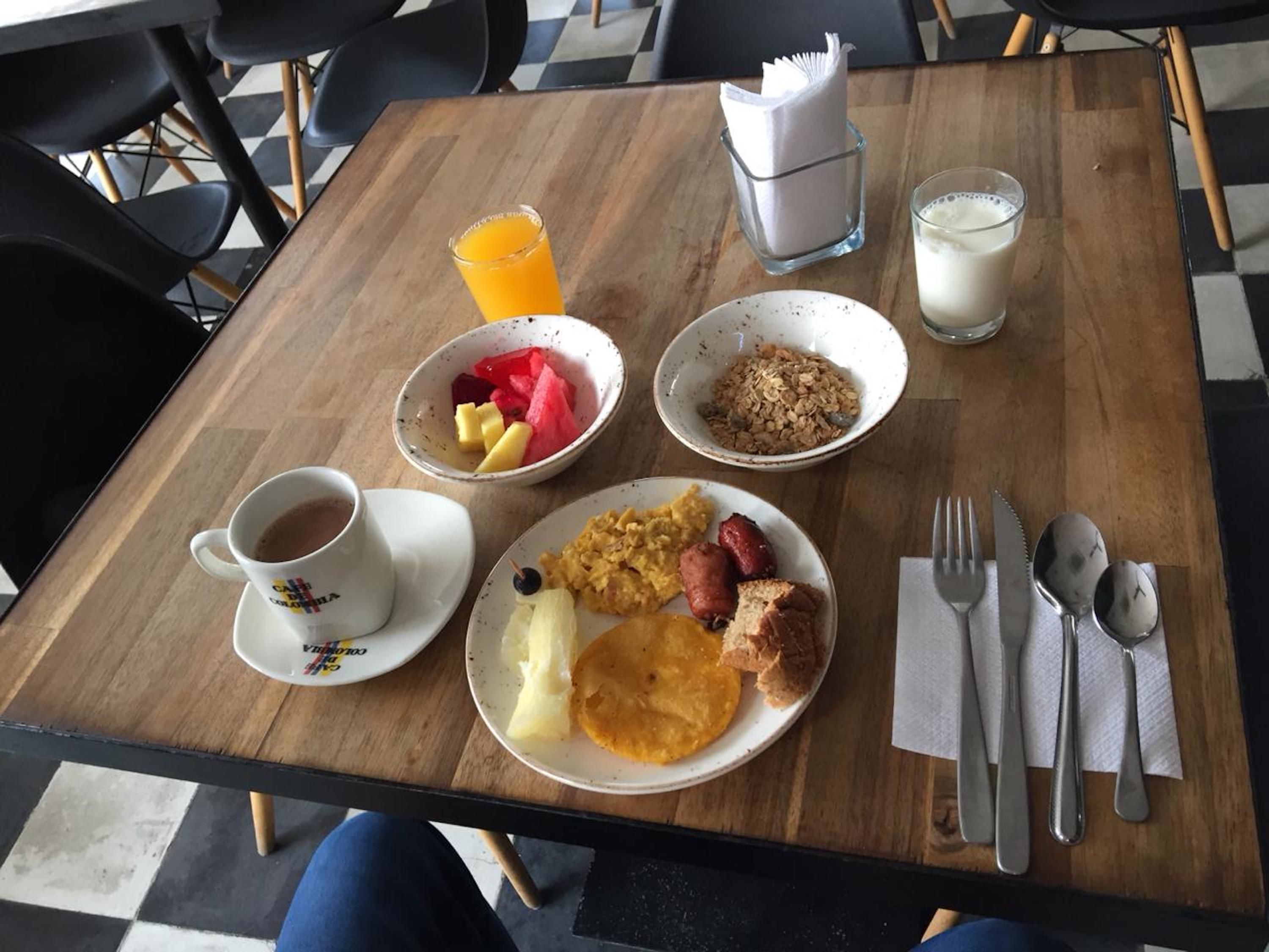 Una mesa de desayuno con una taza de café, un bol de fruta, huevos revueltos, salchichas, tostadas y un vaso de zumo de naranja.