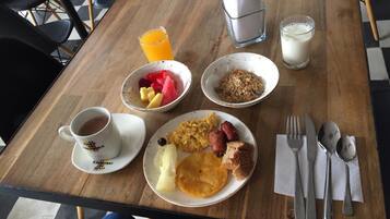 Una mesa de desayuno con una taza de café, un bol de fruta, huevos revueltos, salchichas, tostadas y un vaso de zumo de naranja.