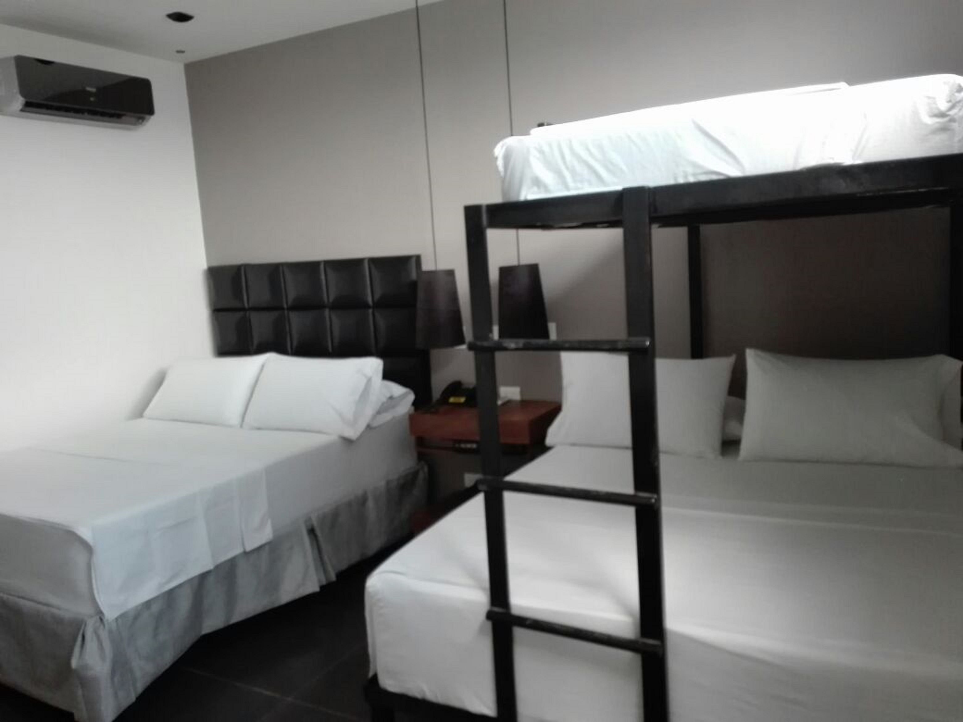 A hotel room with two beds, one with a black headboard and the other with a white headboard. There is a small table with a lamp and a red object on it.