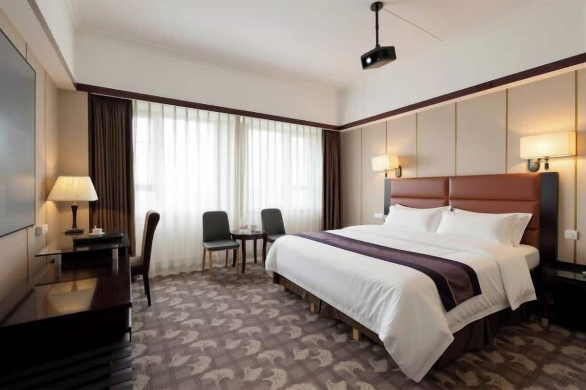 Habitación de hotel con una cama grande, un escritorio con lámpara, dos sillones, una mesita y un amplio ventanal con cortinas.