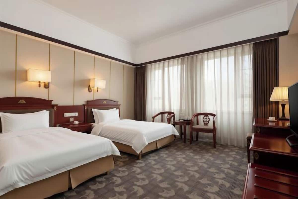 Habitación de hotel con dos camas, un escritorio, una silla y un ventanal con cortinas.
