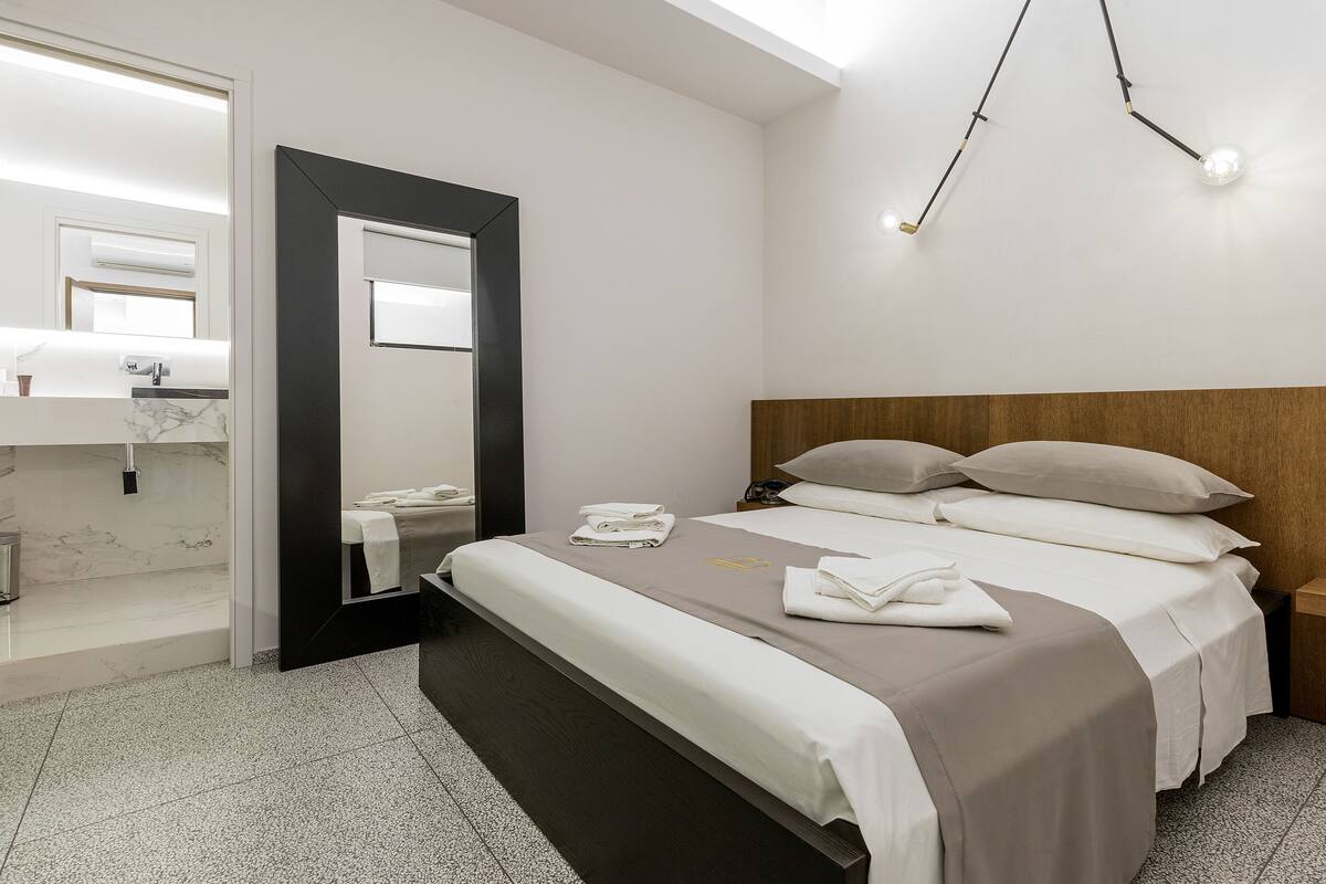 A modern hotel room with a large bed, a wooden headboard, a bedside table, and a mirror.