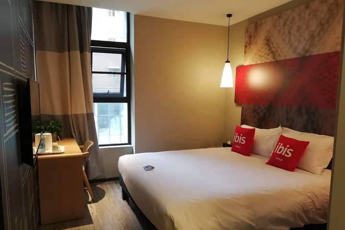 Habitació d'hotel amb llit, tauleta de nit, lamp i finestra amb cortines.