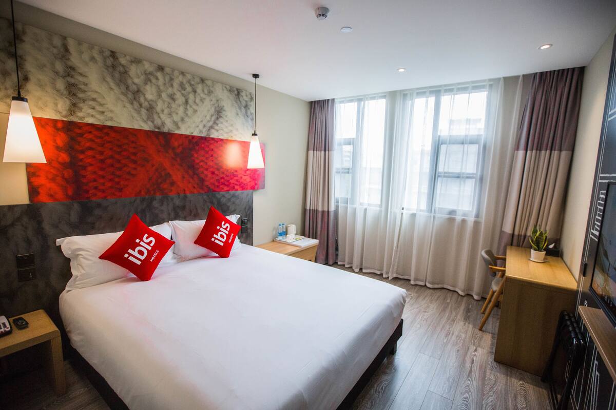Ein Hotelzimmer mit einem großen Bett, zwei roten Kissen, einem Nachttisch, einem Schreibtisch, einem Sessel, einer Zimmerpflanze und einem Fenster mit Vorhängen.