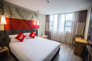 A hotel room with a large bed, two red pillows, a nightstand, a desk, a chair, a potted plant, and a window with curtains. - Ibis Nanchang Bayi Museum Metro Station Hotel Hotel (Nanchang)